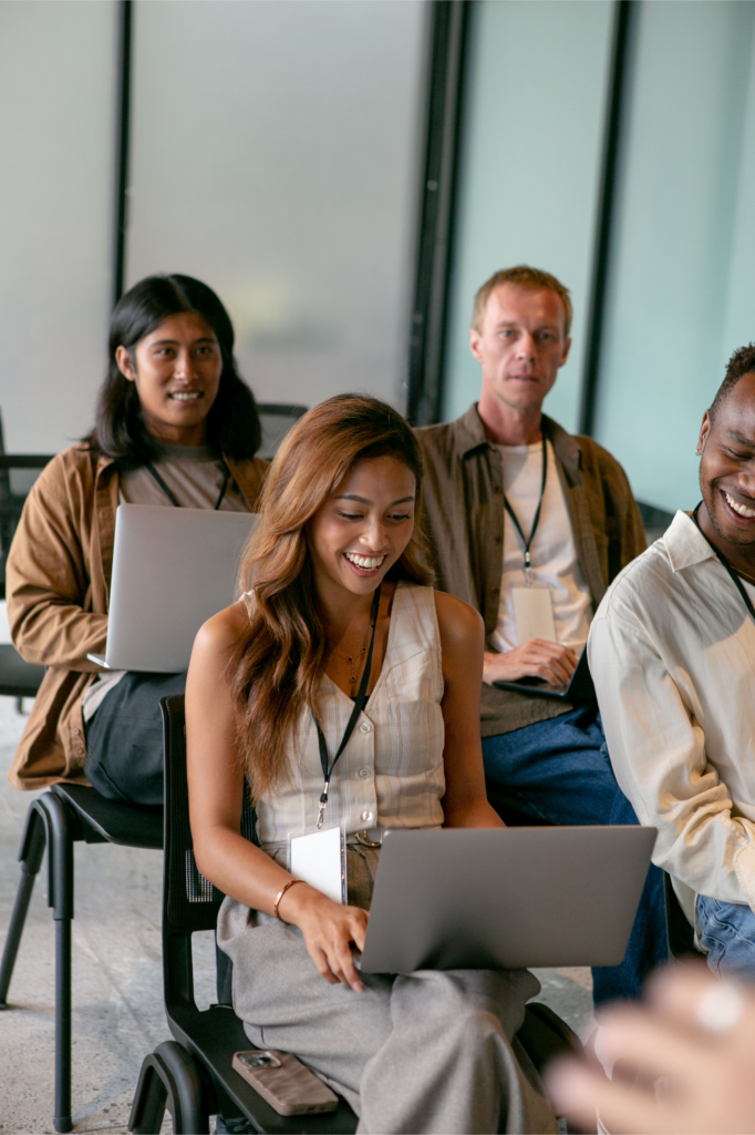 Two men and two women sit in two rows in a conference room. Three of the people have laptops and are smiling. 